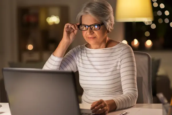 Frau im Rentenalter mit Brille sitzt am Laptop, abendliche Wohnzimmeratmosphäre