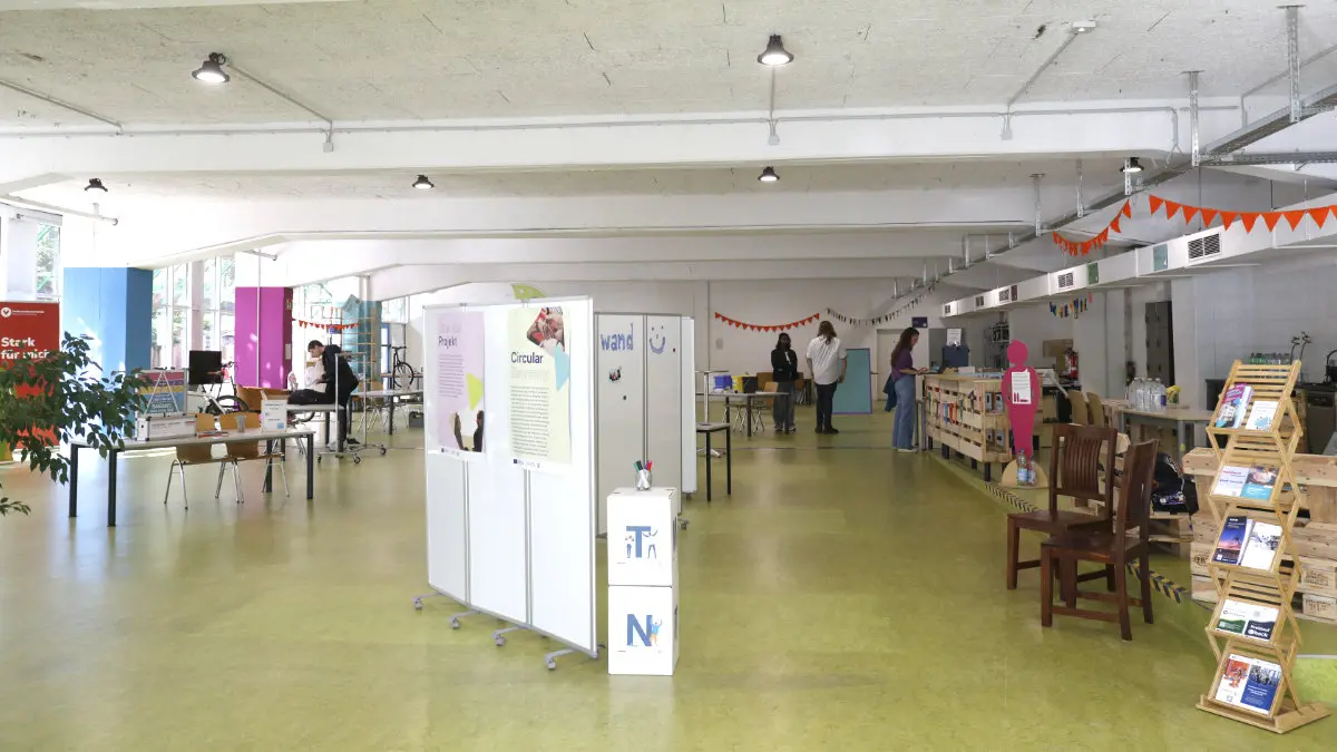 Blick in einen großen Raum, in dem Regale, Info-Aufsteller, Stühle und Tische stehen.