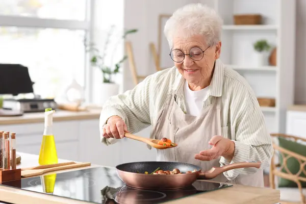 Alte Frau ist in der Küche und bereitet Gemüse in der Pfanne zu, sie lächelt dabei 
