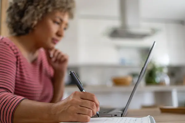 Eine Frau mittleren Alters sitzt in einer Küche am Laptop und macht Notizen auf einem Zettel