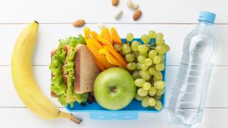 In einer Brotzeitbox liegen ein belegtes Brot, ein grüner Apfel, Paprika-Sticks und grüne Weintrauben. Links daneben liegt eine Banane. Rechts neben der Box liegt eine Flasche mit Wasser.