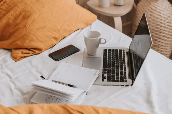Auf einem Bett liegen Laptop, Kalender, Tasse und Handy