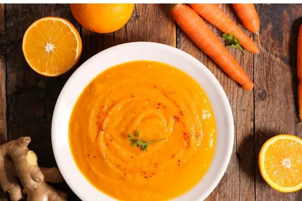 Ein Teller Möhrensuppe, daneben liegen Ingwer, Orangen und Möhren