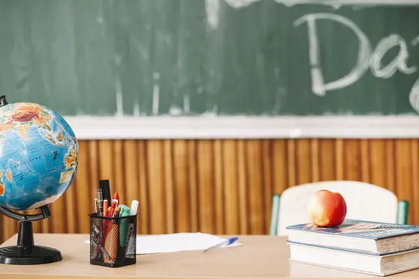 Lehrerpult mit Globus, Stiften, Büchern und einem Apfel vor einer grünen Kreidetafel.