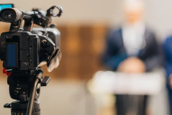 Nahaufnahme einer TV-Kamera, im Hintergrund zwei Personen am Rednerpult