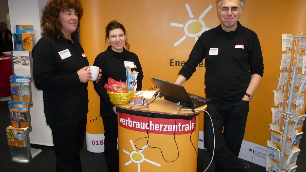 Drei Menschen stehen an einem Informationsstand und lachen in die Kamera.