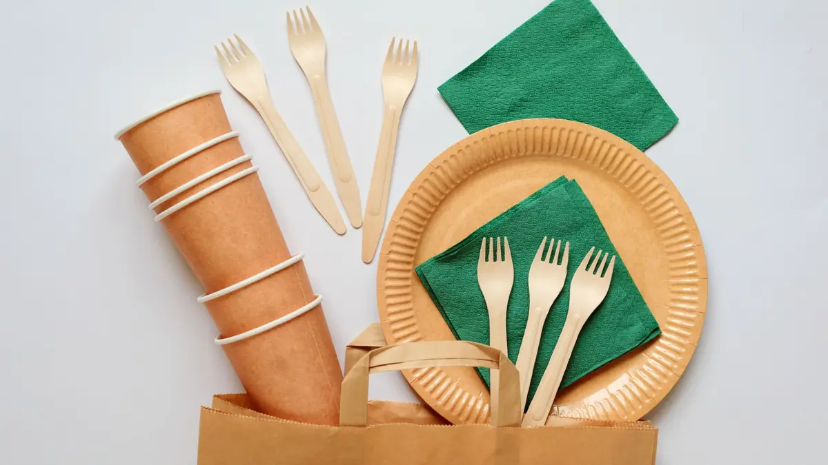 Becher, Teller und Servietten aus Papier und Besteck aus Holz über einer Papiertüte