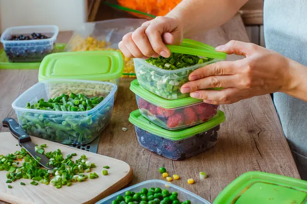 Auf einer Küchenarbeitsplatte befinden sich mehrere Vorratsdosen gefüllt mit frischem Obst und Gemüse. Eine Person schließt eine Dose mit fein geschnittenen Frühlingszwiebeln.