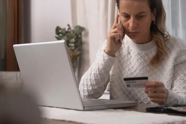 eine Frau sitzt telefonierend vor ihrem Laptop und guckt verunsichert auf ihre Kreditkarte