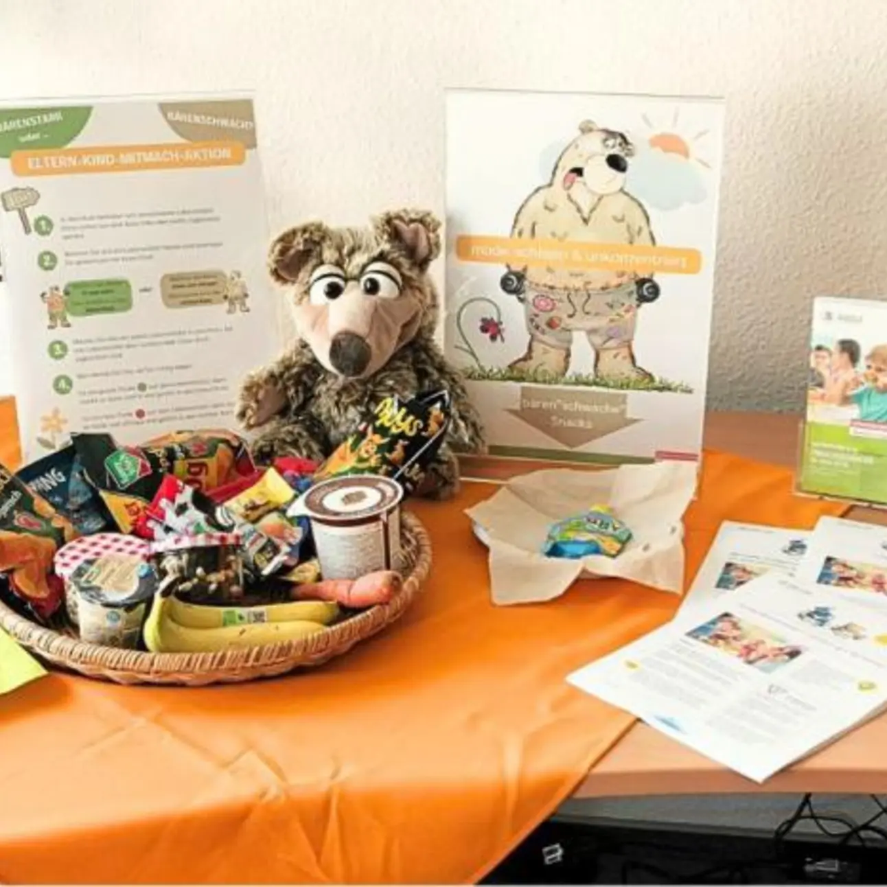 Tisch mit Flockenquetsche, Banane, Möhre, Chipstüte und weiteren Materialien wie Flyer und Aufsteller.