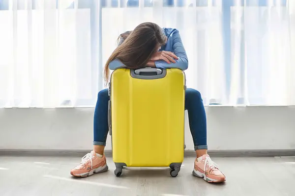 Frau mit gelbem Koffer sitzt am Flughafen. Das Gesicht ist nnicht zu sehen, der Kopf ruht auf dem gelben Koffer.
