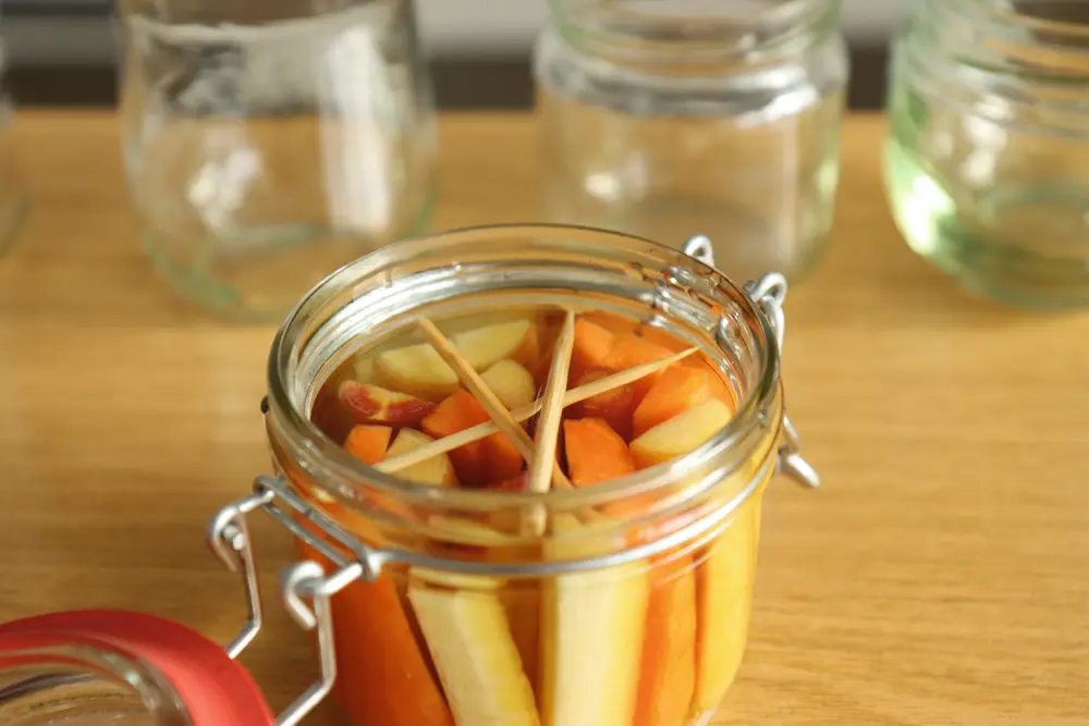 Ein offenes Einmachglas mit Gemüsesticks und Wasser. Holzstäbchen, die zwischen die Glaswände gepresst sind, halten das Gemüse unter der Wasseroberfläche.