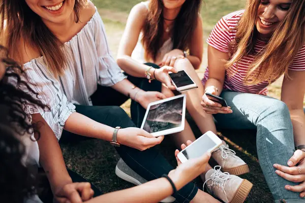 Eine Gruppe junger Frauen. Sie sitzen in einem Park und schauen auf ihre Handys und Tablets.