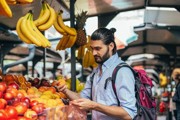 junger Mann mit Bart und Rucksack am Obststand in Markthalle