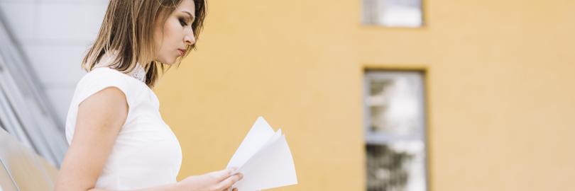 Eine junge Frau steht vor einer Immobilie und sieht Dokumente durch