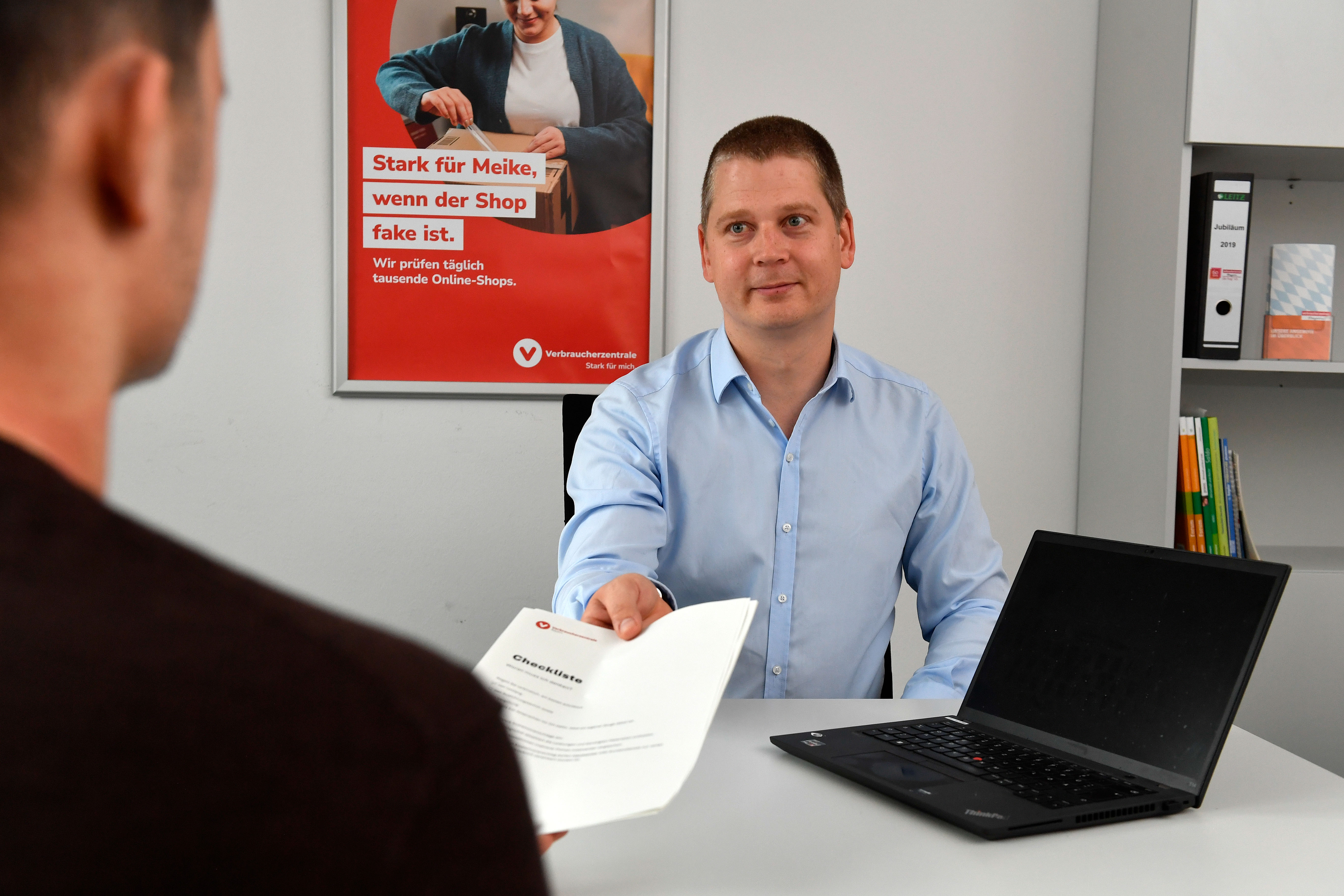 Ein Berater im Gespräch mit einem Verbraucher. Er reicht ihm eine Checkliste mit wichtigen Informationen