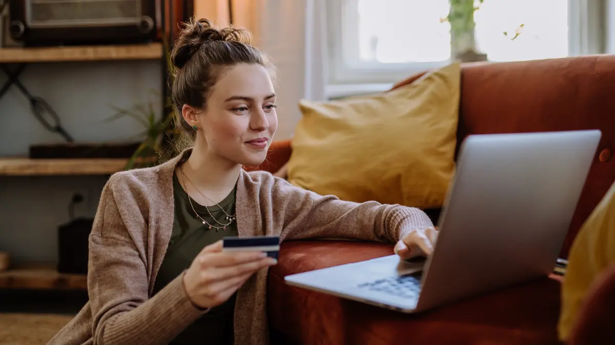Eine Frau sitzt auf dem Boden vor ihrem Laptop, der auf einer ockerfarbenen Couch steht. Eine Hand greift an die Tastatur und mit der anderen Hand hält sie eine Kreditkarte. Sie lächelt.