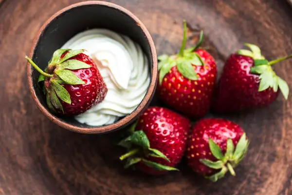 Eine Schüssel mit Sahne steht auf einem Tisch. In der Schüssel liegt eine, neben der Schüssel liegen vier Erdbeeren.