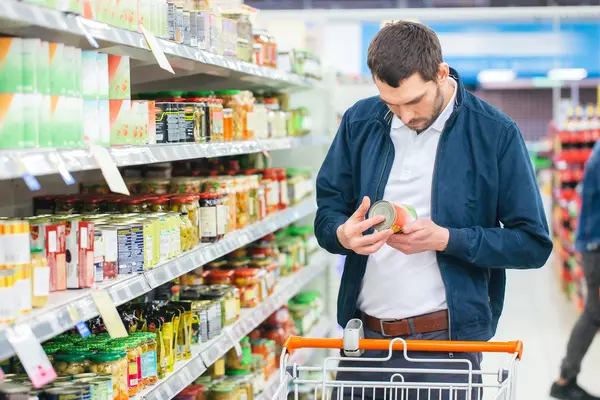 Mann steht mit Einkaufswagen vor einem Supermarktregal und betrachtet die Inhalststoffe eines Produkts