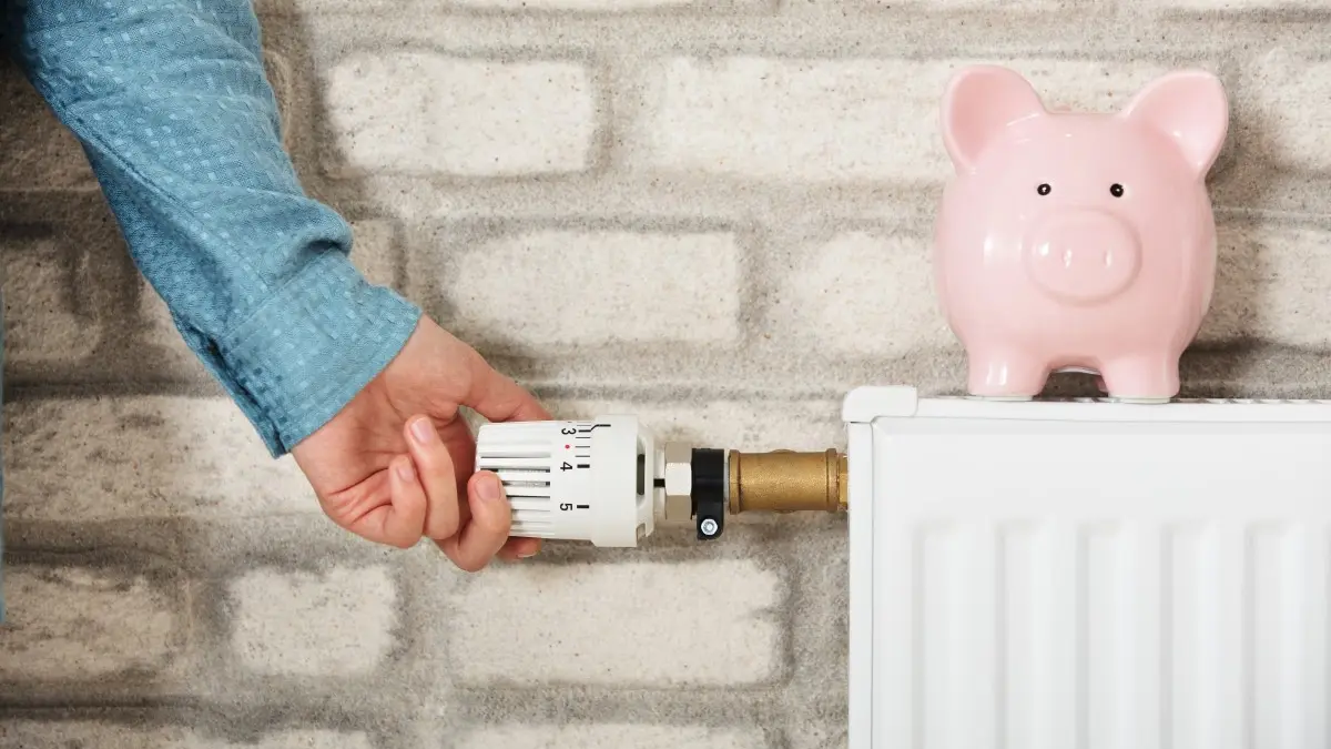Eine Hand dreht an einem Heizungsthermostat, auf dem Heizkörper steht ein Sparschwein.