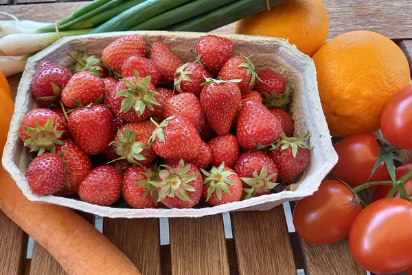 Erdbeeren in einer Pappschale stehen auf einem Holztisch. Daneben liegt anderes Obst und Gemüse.