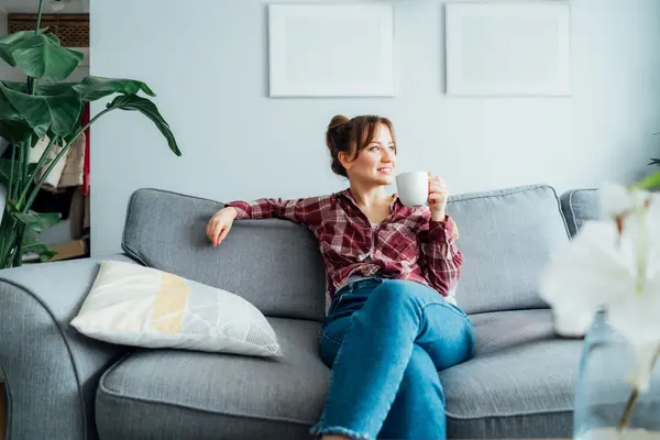Eine Frau sitz entspannt mit einer Tasse in der Hand auf dem Sofa