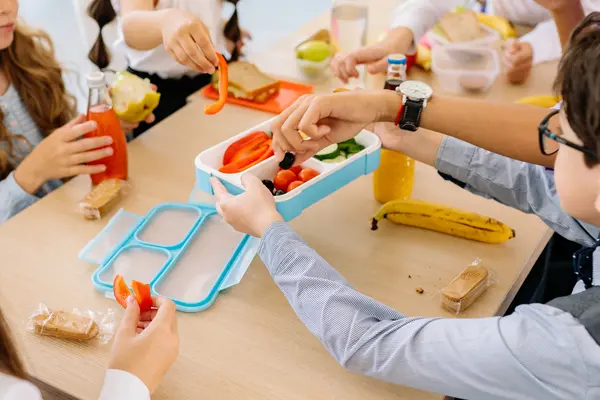 An einem Tisch sitzen 4 Kinder. Alle greifen nach einer hellblauen Brotbox, in der sich verschiedenes Gemüse befindet. Auf dem Tisch sind Lebensmittel, wie belegte Brote sowie Obst und Gemüse verteilt. In Glasflaschen mit verschiedenen Säften und Wasser stehen auch auf dem Tisch.  
