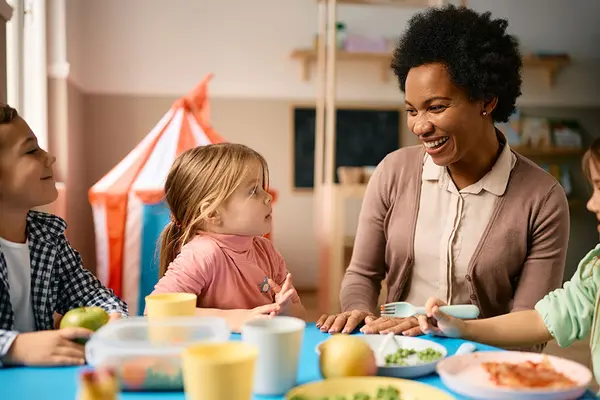 Eine Erzieherin sitzt mit Kindern im Kindergartenalter am Esstisch