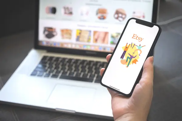 Eine Hand hält ein Handy, zu sehen die Etsy-App, im Hintergrund ein Laptop mit dem Etsy-Online Shop