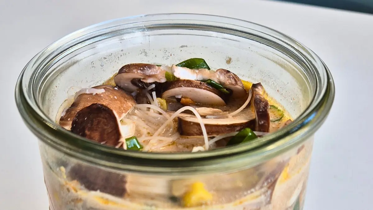 Ein Einmachglas mit fertigem Healthy Homemade Ramen: Reisnudeln, Pilze, Maiskörner und Frühlingszwiebeln in einer würzigen Brühe. Das Glas steht auf einer weißen Oberfläche