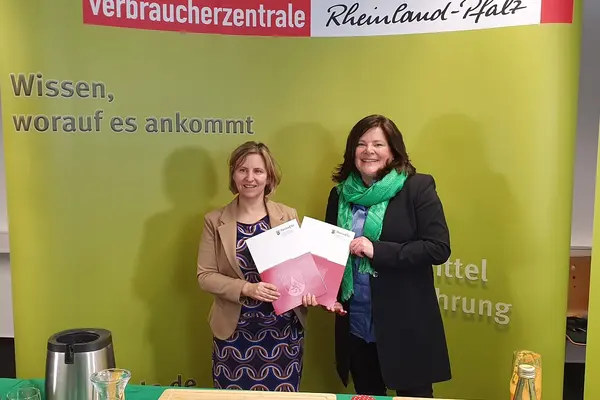 Zwei Frauen vor eienr grünen Wand. Sie halten zwei Dokumentenmappen in die Kamera.