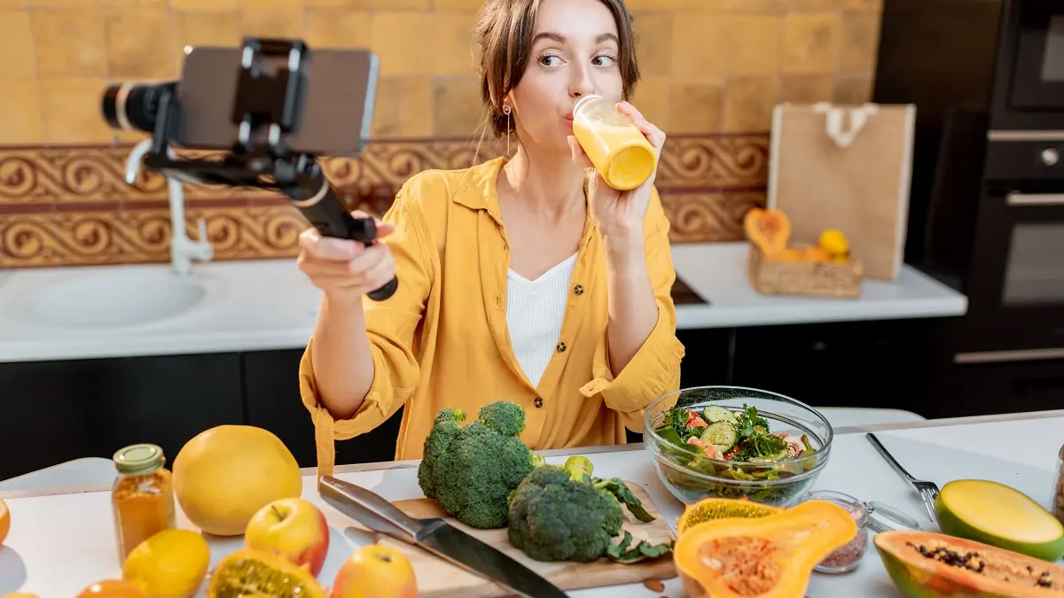 Frau mit Smoothie ind er einen Hand udn Smartphone in der anderen. Sie filmt sich beim Trinken. Vor ihr ist ein Schneidebrett und einiges an Obst und Gemüse