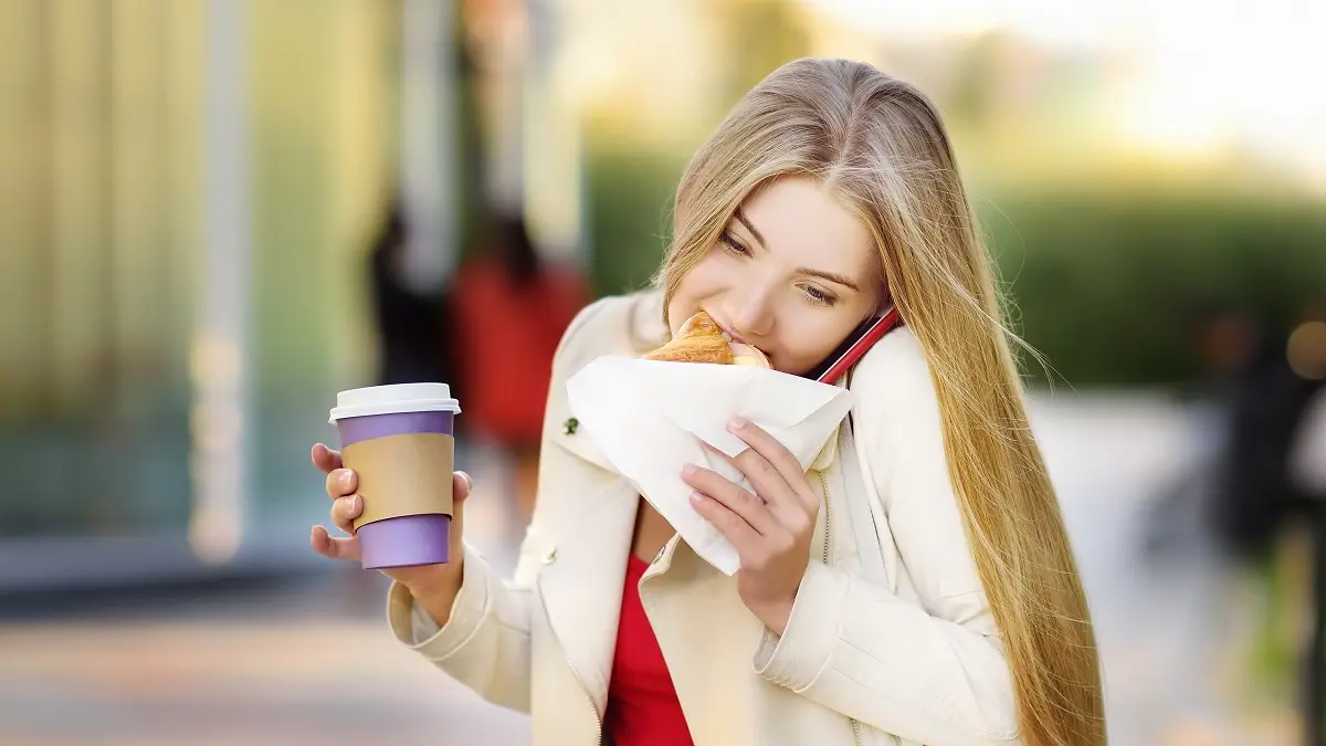 Eine Frau ist unterwegs und beißt in ein Sandwich. Inder anderen Hand hat sei einen Coffee to go in der Hand