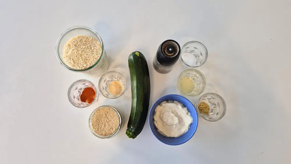 Zutaten für Crispy Zucchini mit Honig-Senf-Dip auf einem weißen Hintergrund: eine frische Zucchini, Schälchen mit Pankobröseln, Parmesan (oder Hefeflocken), Paprikapulver (edelsüß), Oregano, Salz und Pfeffer, eine Schale mit griechischem Joghurt, sowie kleine Gläser mit Olivenöl, Honig, Senf und Zitronensaft. Alles bereit für die Zubereitung.