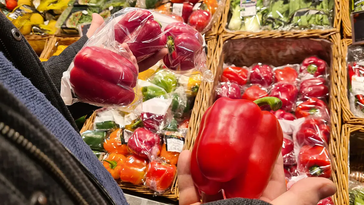 Im Supermarkt hält jemand unverpackte und in Plastikfolie verpackte rote Paprika in der Hand