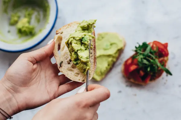 Frauenhände bestreichen ein Stück Ciabatta mit grüner Creme