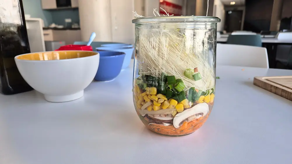 Ein Einmachglas auf einer weißen Arbeitsplatte, gefüllt mit den Zutaten für Healthy Homemade Ramen: Schichten aus Reisnudeln, geraspelter Möhre, Maiskörnern, geschnittenen Frühlingszwiebeln und Pilzscheiben. Im Hintergrund stehen eine weiße Schüssel, eine blaue Schüssel und Küchenutensilien. Das Glas ist bereit für die Meal-Prep-Aufbewahrung.