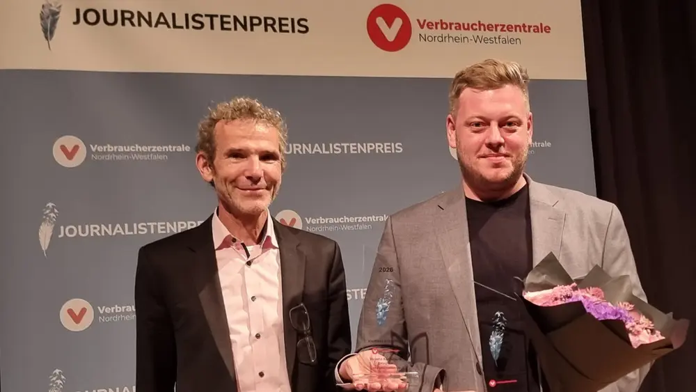 Zwei Männer stehen nebeneinander und lächeln in die Kamera. Einer hält einen Blumenstrauß und eine Trophäe in den Händen.