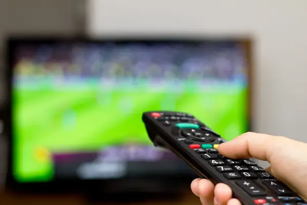 Foto von einer Hand, die eine Fernbedienung hält und auf einen Fernsehbildschirm richtet, auf dem ein Fußballspiel läuft