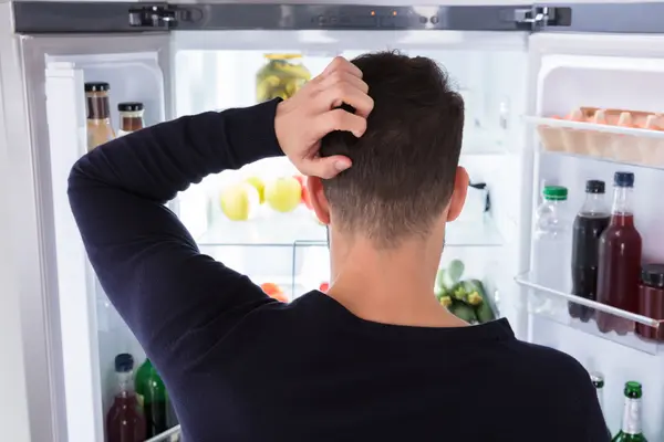 Ein Mann guckt in einen geöffneten Kühlschrank und kratzt sich dabei am Kopf.