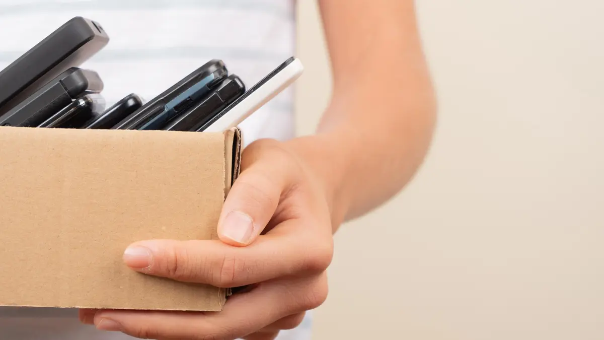Nahaufnahme einer Person, die einen kleinen Pappkarton hält, der mit mehreren alten Mobiltelefonen und Smartphones gefüllt ist.