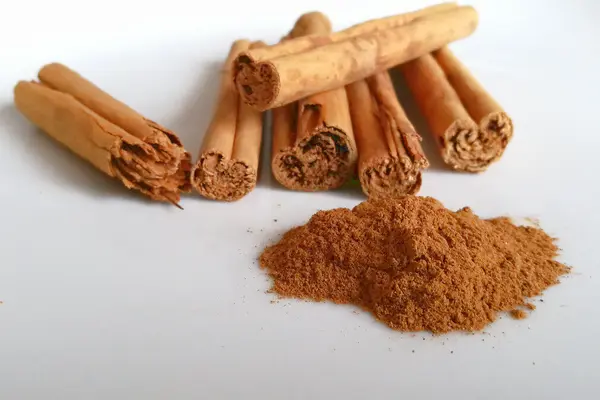 Sechs Cassia-Zimtstangen liegen auf einer weißen Fläche; davor ein Häufchen gemahlener Zimt