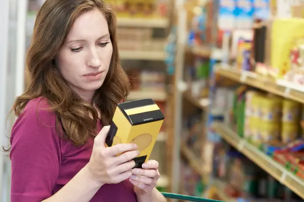 Eine Frau steht vor dem Supermarktregal, hält eine Verpackung in der Hand und schaut sie sich an.