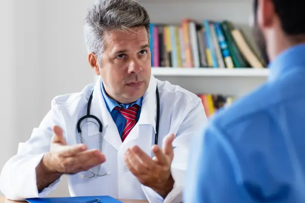 Ein Hausarzt sitzt am Tisch und spricht mit einem Patienten, von dem das Bild nur den Rücken zeigt.