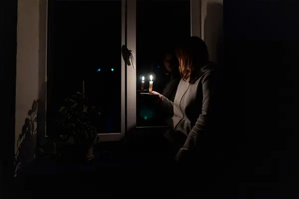 Frau mit brennender Kerze in der Hand steht in dunklem Raum am Fenster