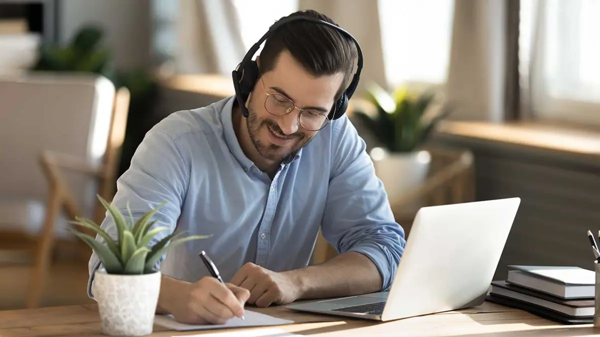 Videoberatung: Ein Mann sitzt mit Laptop und Headset an einem Tisch und macht Notizen