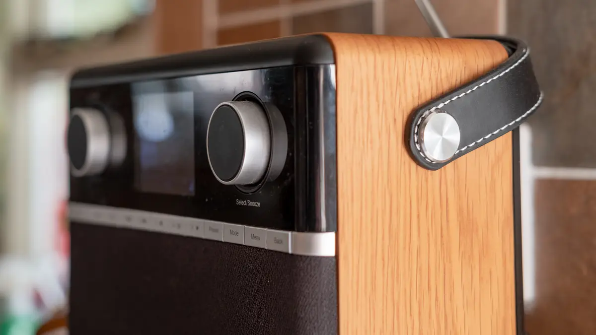 Nahaufnahme eines Digitalradios mit Holzgehäuse, schwarzer Frontblende, silbernen Drehreglern, kleinem Display, Bedientasten (Preset, Mode, Menu, Back) sowie schwarzem Ledertragegriff und ausgezogener Teleskopantenne, im Hintergrund unscharfe Fliesen.