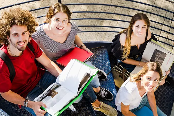 Vier junge Erwachsene sitzen mit Büchern auf einer Wendeltreppe und schauen nach oben in die Kamera