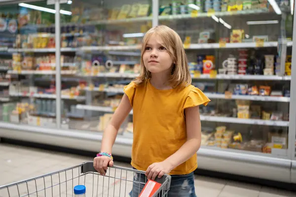 Ein Kind in einem gelben Shirt steht neben einem Einkaufswagen im Supermarkt