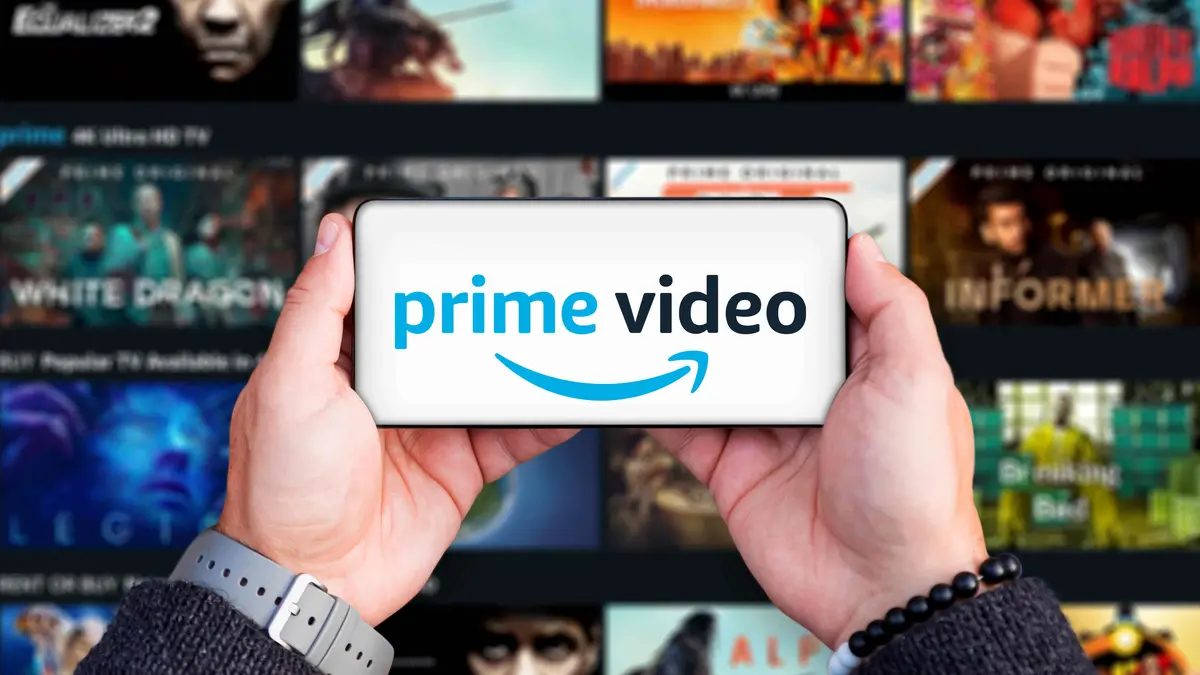 Händ ehalten Handy auf dem der Schriftzug prime Video zu sehen ist. Im Hintergrund sieht man den die Prime Fimauswahl auf einem großen Fernseher